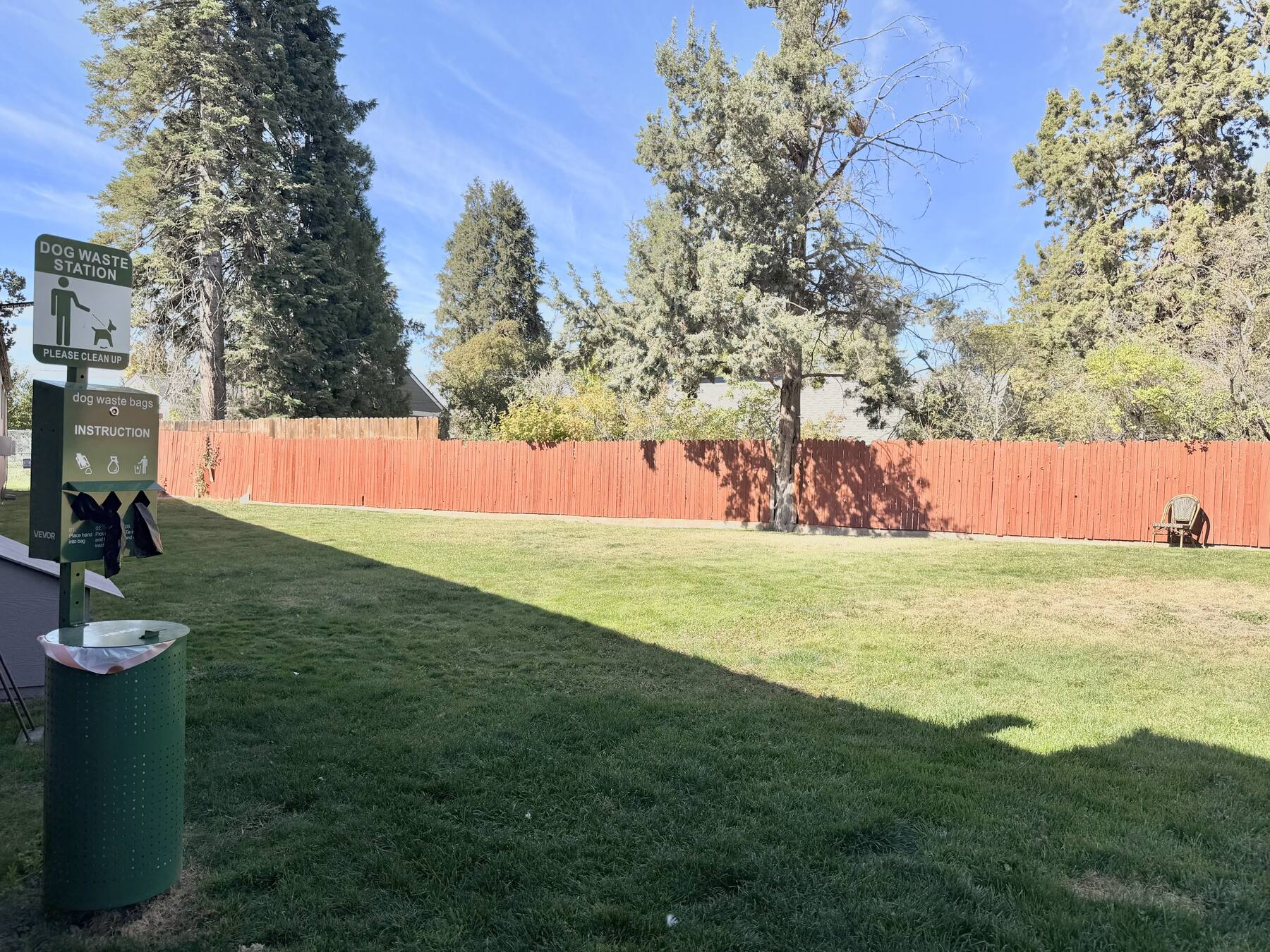 Fenced grassy pet area with a dog waste station, shaded by pines