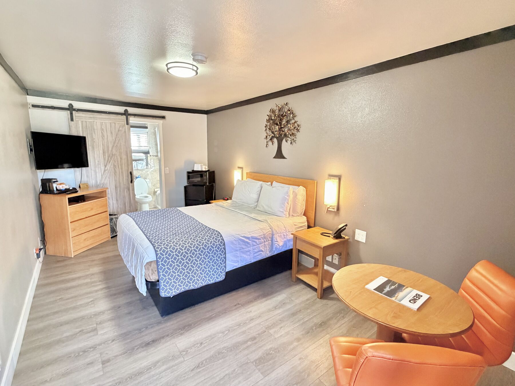 Cascade Queen room with a queen bed between wall sconces, tree wall art, a dresser with TV, a barn-door bathroom, and a round table with orange chairs