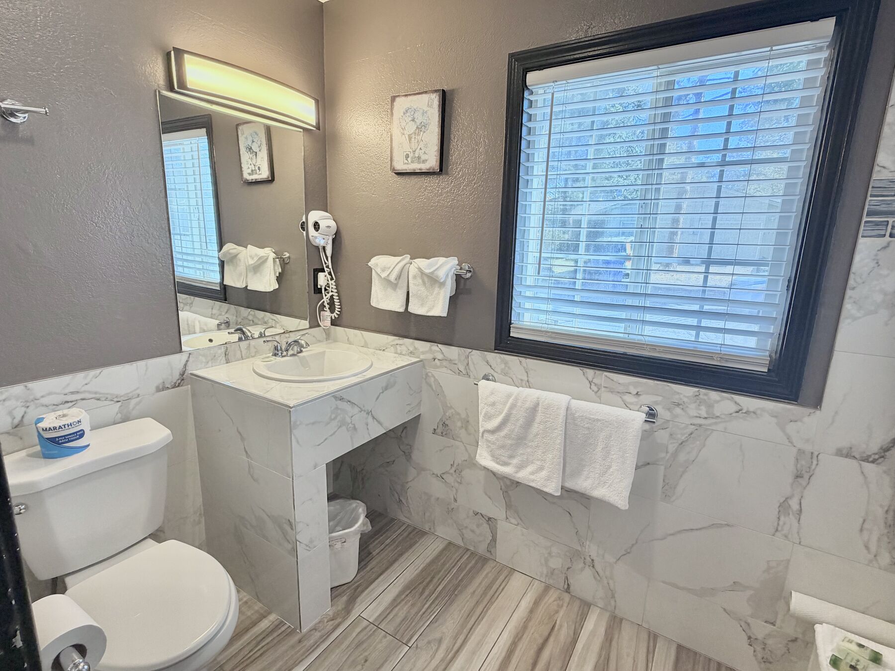 Cascade Queen bathroom with a marble-tiled vanity, lit mirror, hair dryer, fresh towels, and a large window with blinds